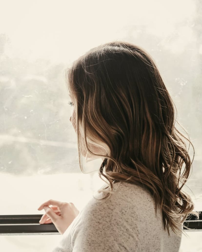 A woman stands at a bright window, resting her hand on the sill and looking out.