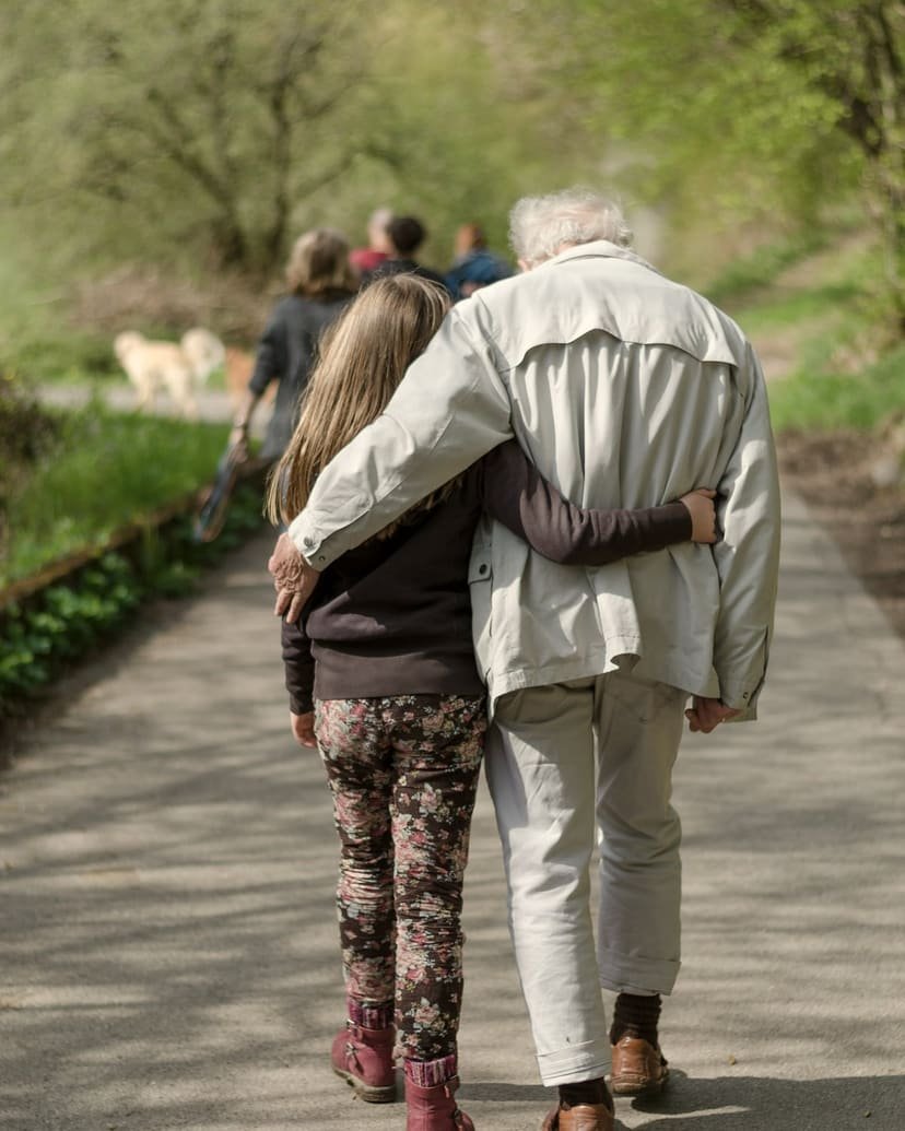 An older adult and a young person walk side by side along a leafy path, arms around each other.