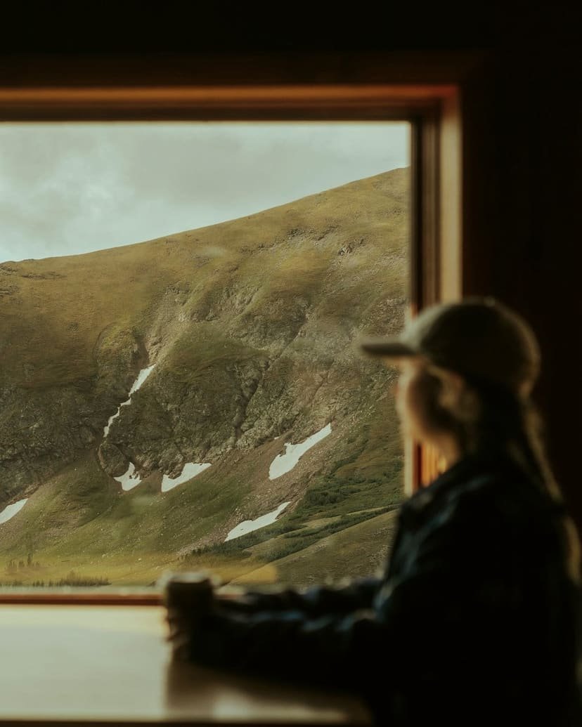A person in a cap sits at a desk beside a wide window overlooking green hills.