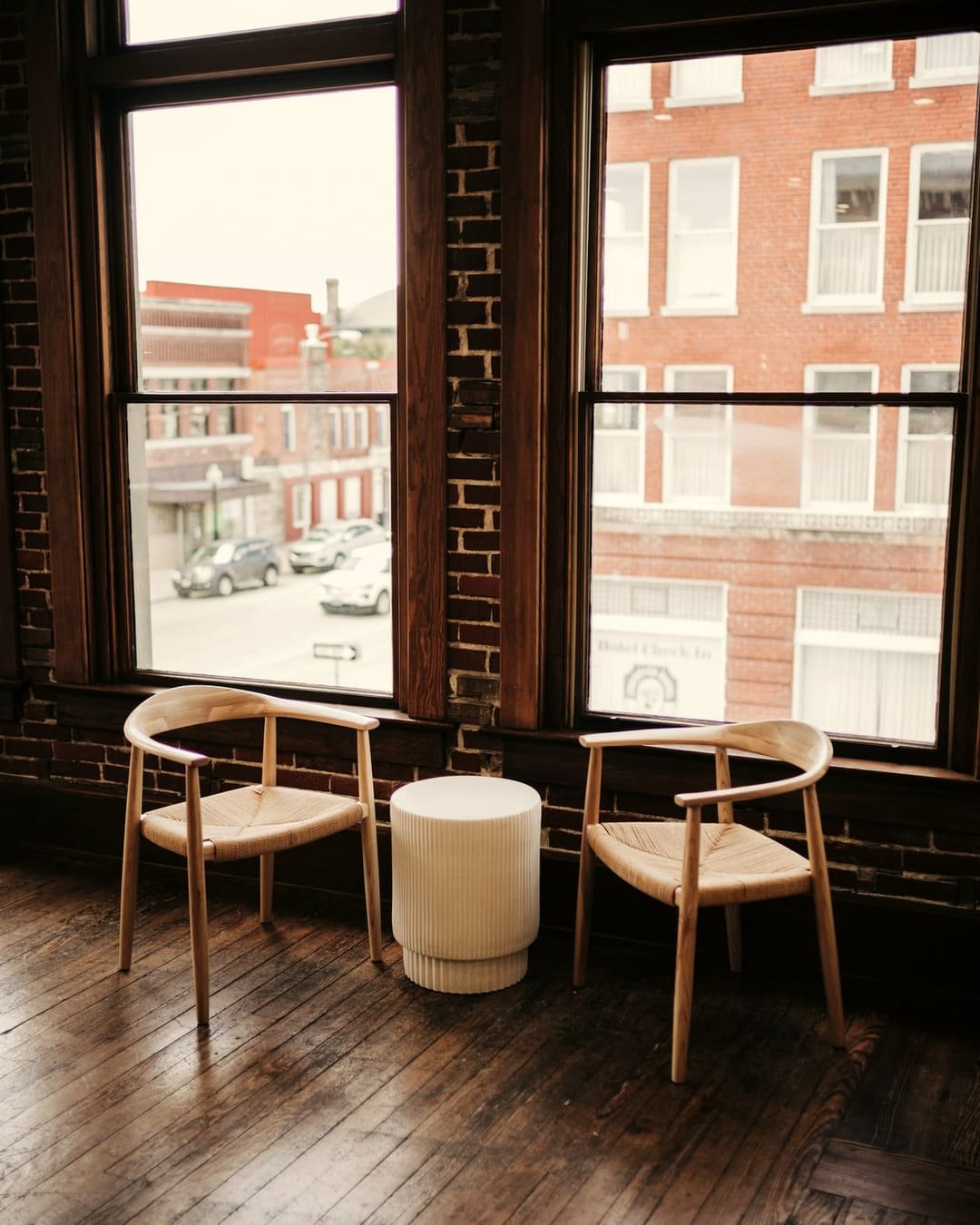 Two wooden armchairs and a small side table beside tall windows in a quiet, light-filled room.